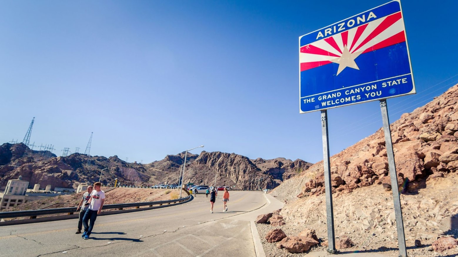 People walking on a road next to an Arizona state welcome sign in the daytime.