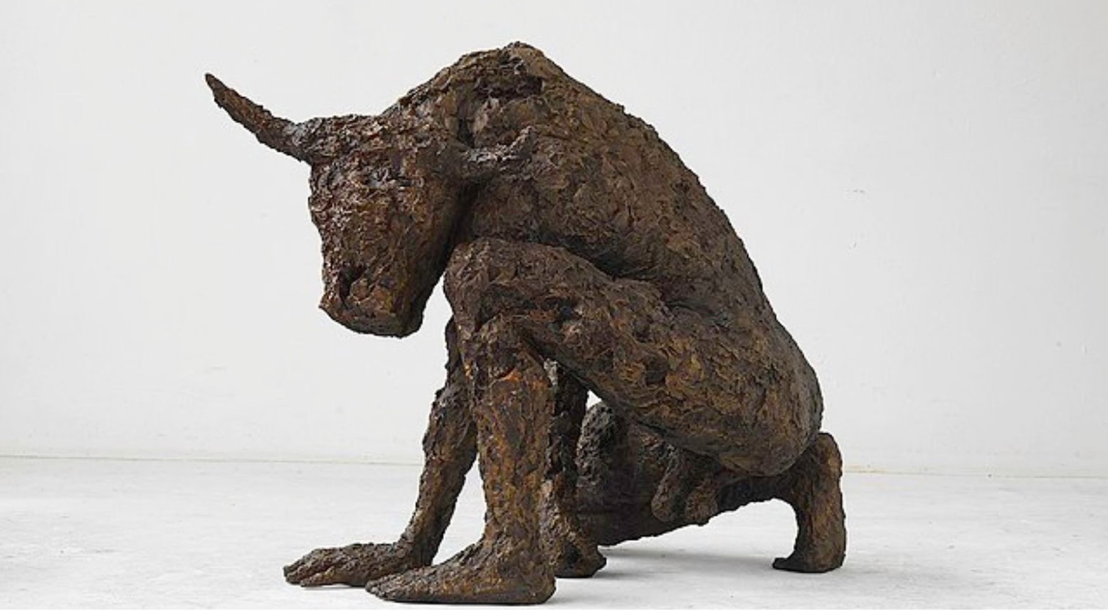 A bronze statue of a minotaur. The statue has a bull's head and a human body. The statue is on one knee, with both hands on the floor.
