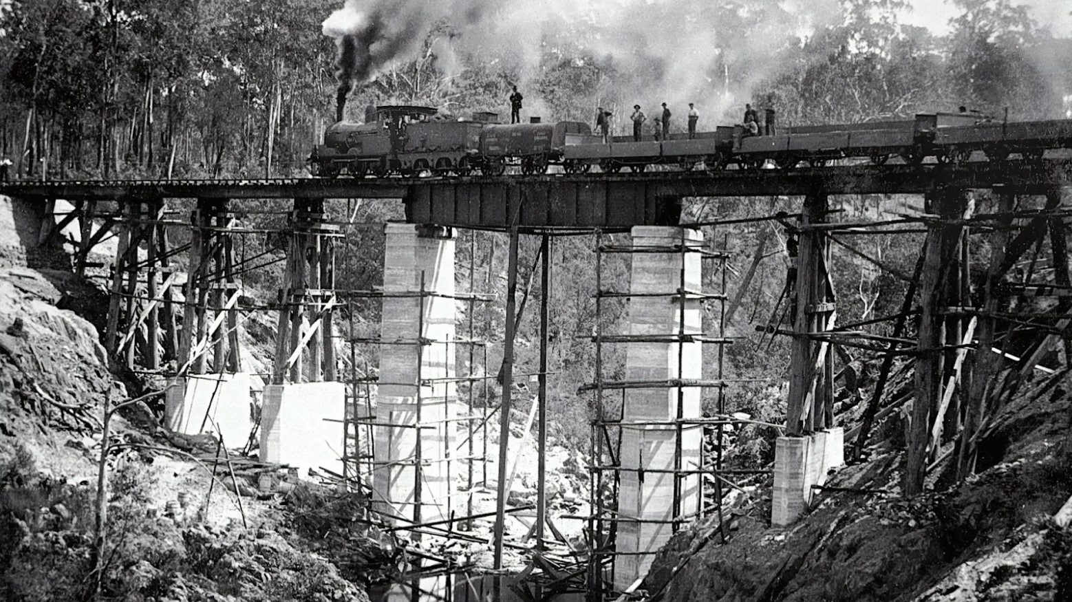This is a black-and-white image of some workers working on a railway line. The train is on a railway track, and workers are standing on top of it. Scaffolding leads up to the track.