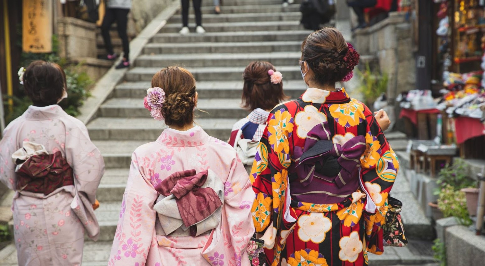 The geisha district. There are four geishas in floral kimonos standing at the foot of some steps. Some people are walking down the steps.