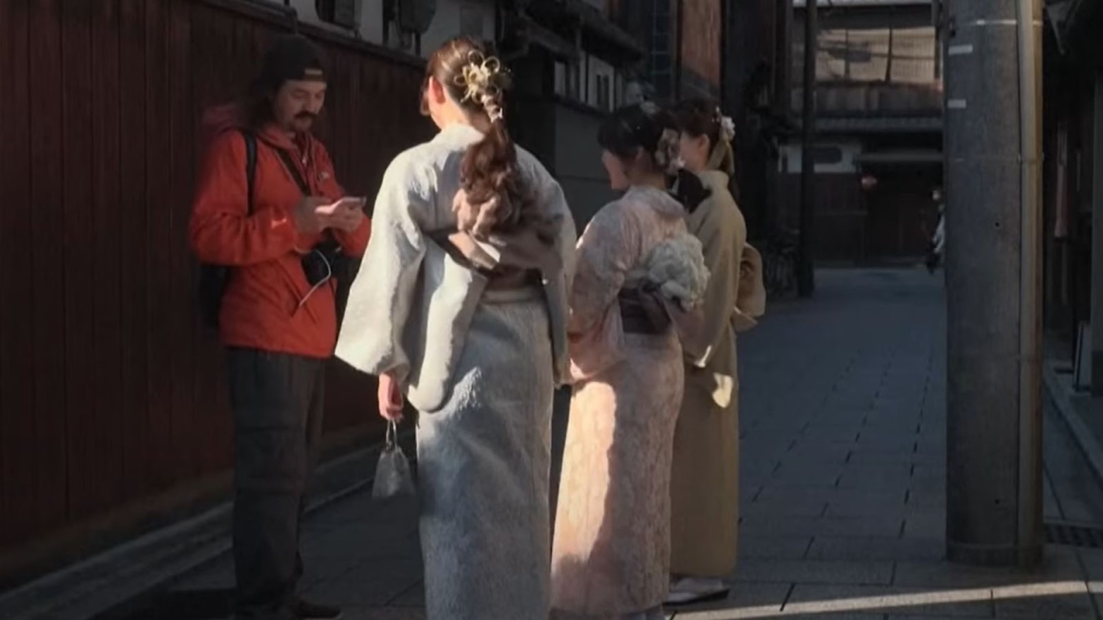 A tourist in an orange coat standing looking at his phone. Standing in front of him are three geishas, one in a blue kimono, one in a pink kimono, and one in a yellow kimono.
