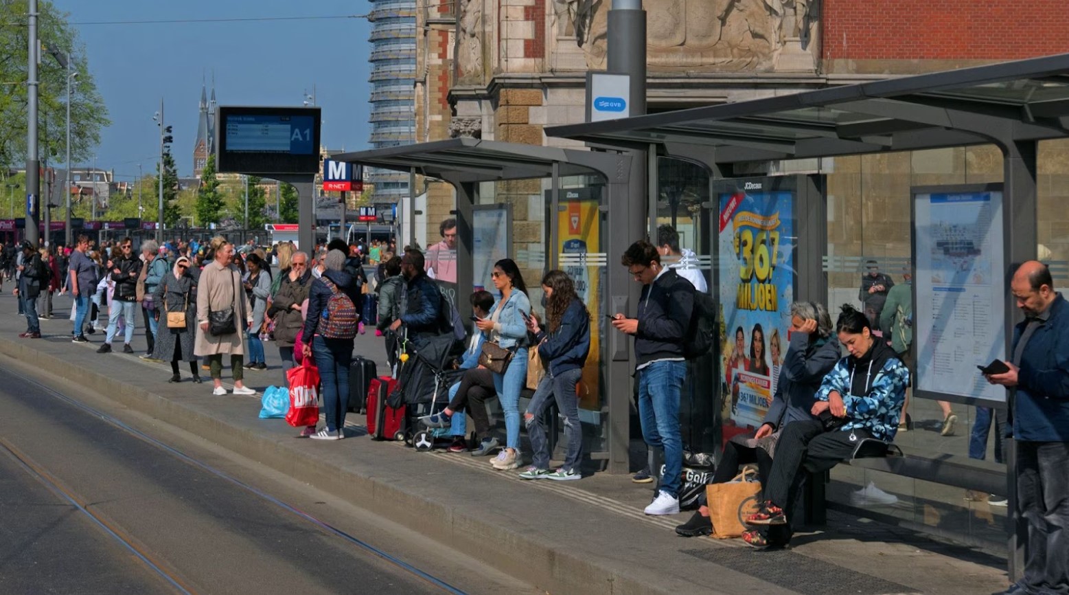 A crowd of people standing at a bus stop. Some people are sitting in the bus shelter.