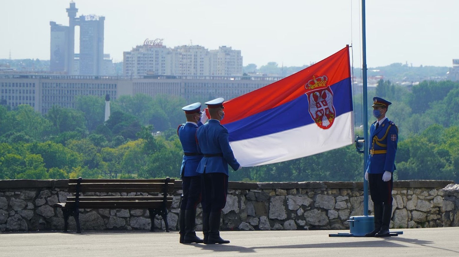 Three soldiers raising the Russian flag on a flagpole. Behind them is a view consisting of green trees and high-rise buildings.