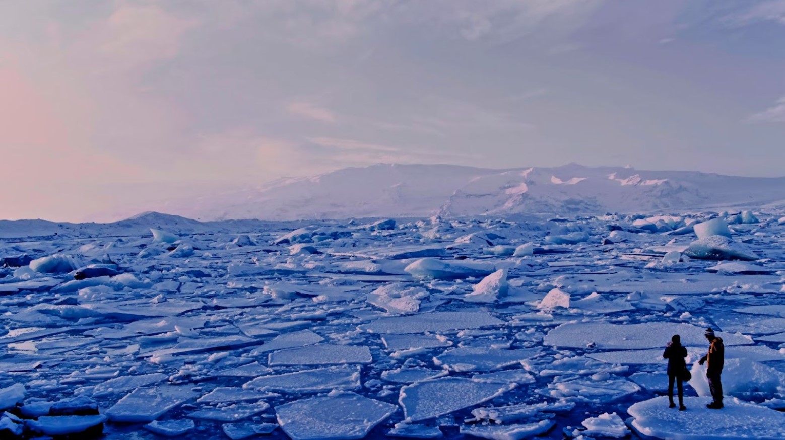 An image of broken sheets of ice in the sea. Two people are standing on one of the ice sheets.