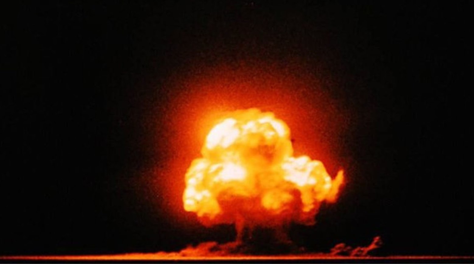 A color photo of the first nuclear test explosion from the Manhattan Project. The background is black, and the explosions consist of a cloud of red, orange, and yellow.